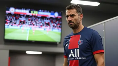 A 35-year-old Paris FC assistant coach, wearing a plain unmarked blue and red jersey made of high-quality cotton, stands...