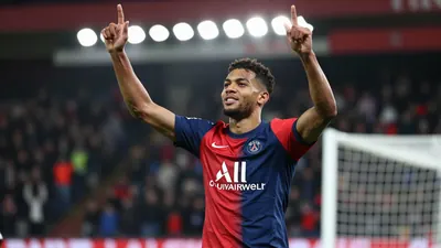 A young, athletic striker in a red and blue Paris FC jersey, mid-celebration after scoring a goal, arms raised, face lit...
