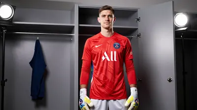 A 24-year-old Paris FC goalkeeper, wearing a red jersey and white shorts, stands in front of a locker room door, looking...