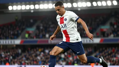 A 21-year-old Paris FC winger, wearing a white jersey with a blue stripe, a blue short, and blue socks, with a black...