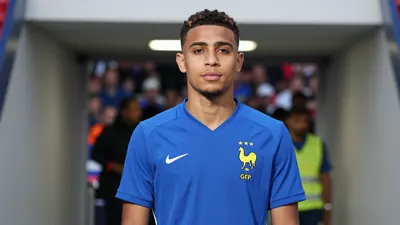 A 26-year-old French forward in a clean solid blue football kit, standing in a stadium tunnel with blurred crowd in the...